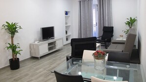 Un salon moderne avec une table en verre, des chaises noires, un téléviseur à écran plat, un meuble blanc et deux plantes en pot.