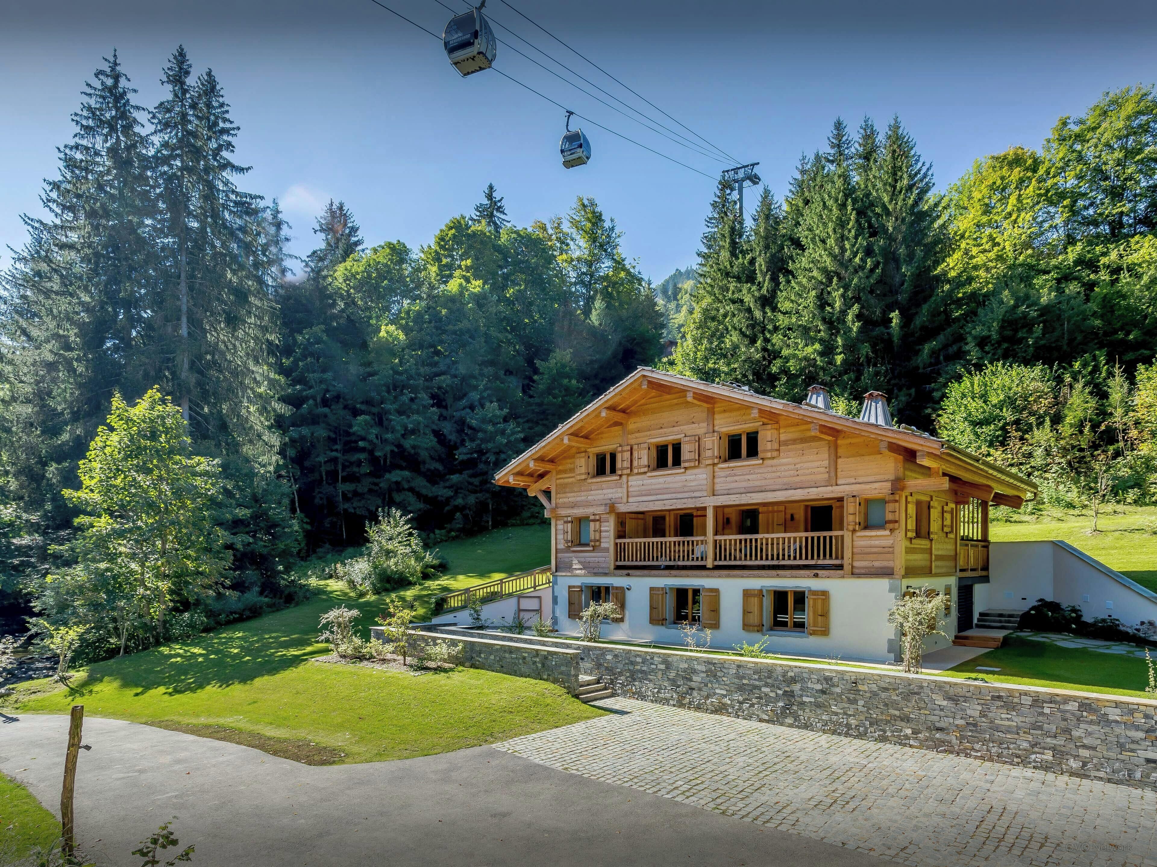 Un chalet en bois doté d’un balcon, entouré d’un mur de pierre et d’une végétation luxuriante.