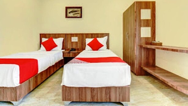 Habitación de hotel con dos camas individuales, cada una con almohada roja y ropa de cama blanca. Hay una mesita de noche de madera entre las camas y un armario de madera apoyado en la pared.