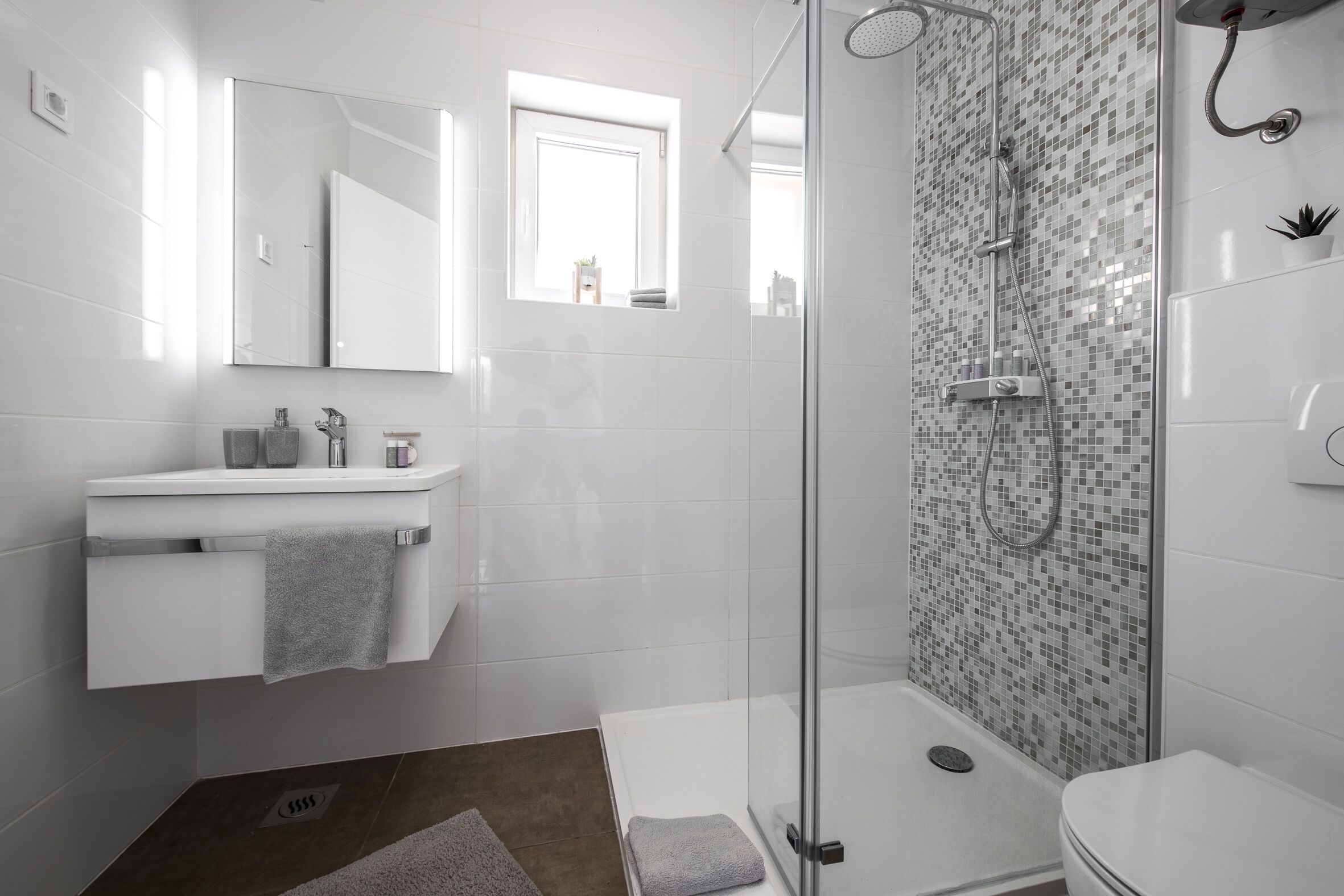A modern bathroom with a glass-enclosed shower, white tiles, a large mirror, and a small window.