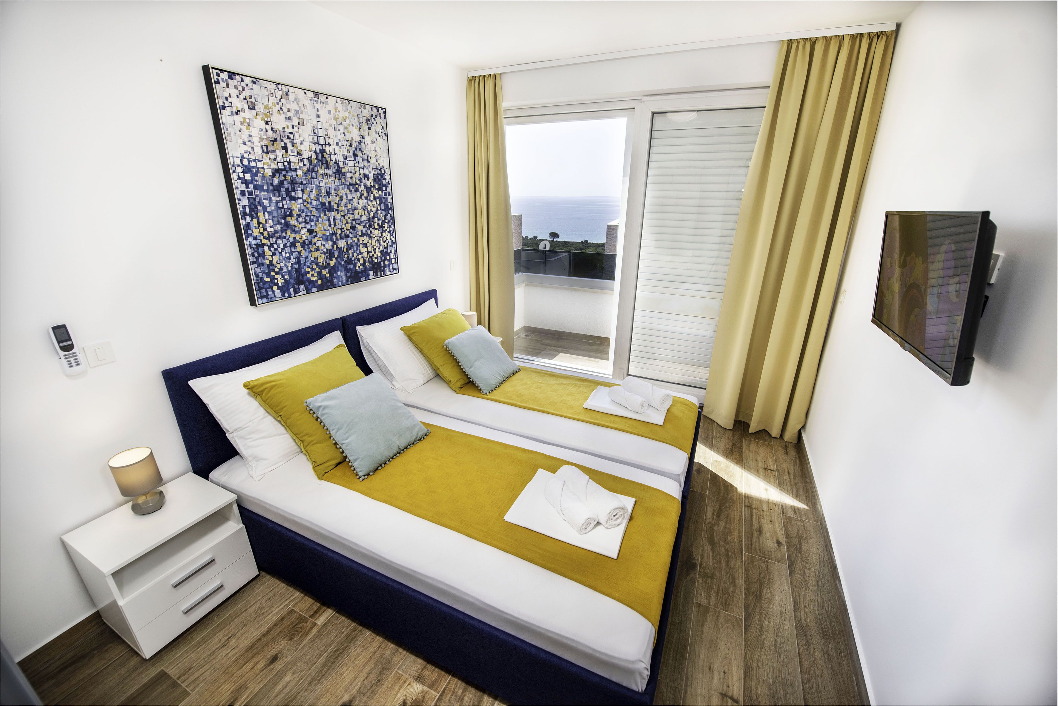 Una cama doble con una colcha color mostaza, dos almohadas y una mesita de noche blanca con una lámpara. Un cuadro está colgado en la pared sobre la cama, y un televisor está montado en la pared a la derecha.