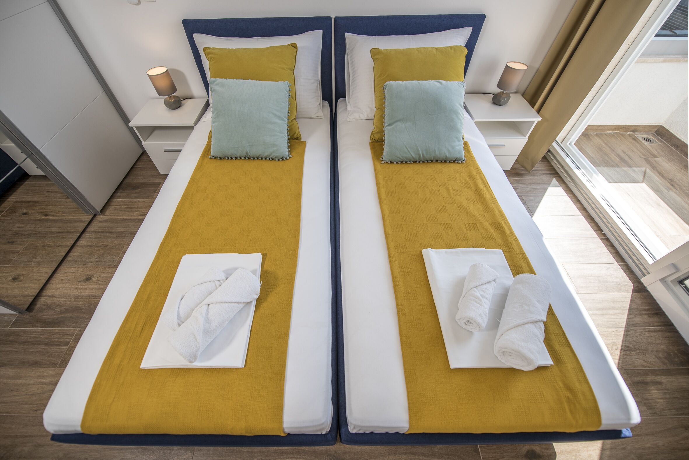 A twin room with two beds, each with a yellow bedspread, white pillows, and a white towel. There are bedside tables with lamps and a window with a view.