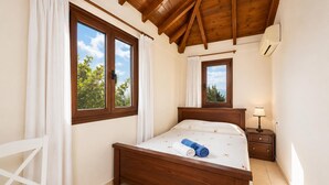 A bedroom with a wooden bed, two windows with curtains, a bedside table with a lamp, and a white chair. - Ekklisies Tria Large Private Pool, A/C, WiFi (Apokoronos)