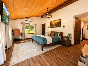 A bedroom with a wooden ceiling, a bed with multiple pillows, a nightstand, a desk with a chair, a window with curtains, and a painting on the wall.