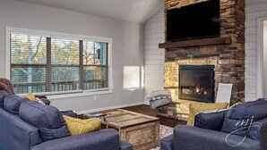 A living room with a stone fireplace, a large window with blinds, and a flat-screen TV mounted above the fireplace. - Norfork Lake: Family Retreat  (Calico Rock)