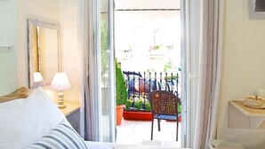 Habitación de hotel con una cama, una mesita de noche con lámpara, un sillón y vistas al balcón exterior.