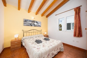Um quarto com cama, mesas de cabeceira, um quadro, uma janela e cortina.