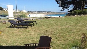 A patio area with outdoor furniture, including chairs and a table, overlooking a grassy area with a view of the sea and a distant shoreline.