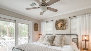 Ein Schlafzimmer mit einem Bett, einem Deckenventilator, einem Nachttisch mit Lampe und Blick ins Freie durch eine Schiebetür.