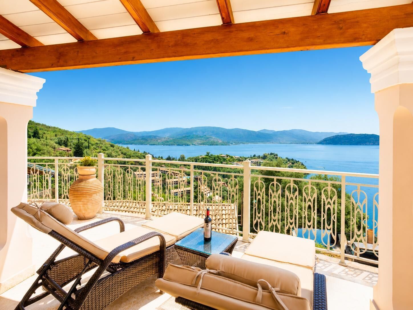Uma área de estar coberta ao ar livre com móveis de vime, uma mesa e vista para um lago e montanhas.