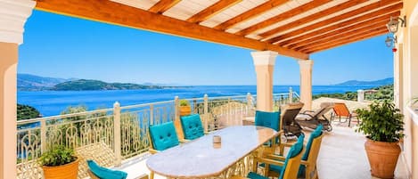 Un espacio de estar al aire libre cubierto, con una mesa y sillas, que ofrece una vista panorámica del mar y las colinas.