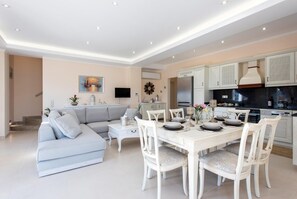 A modern living room with a sofa, dining table, and chairs. There is a kitchen area with cabinets and appliances. - Villa Frosso - Three Bedroom Villa, Sleeps 6 (Kalami)