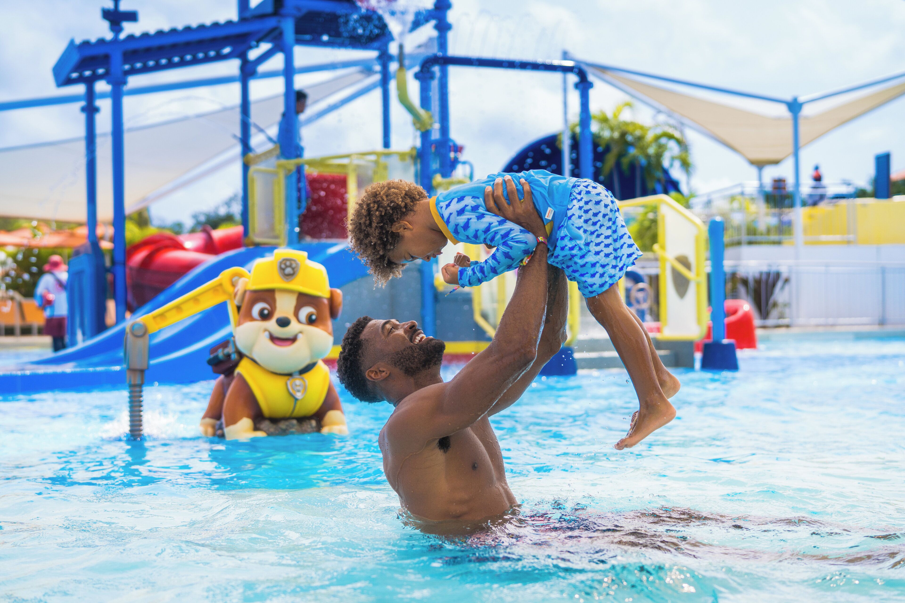 A man holding a child in a swimming pool with a water slide and play structures in the background.