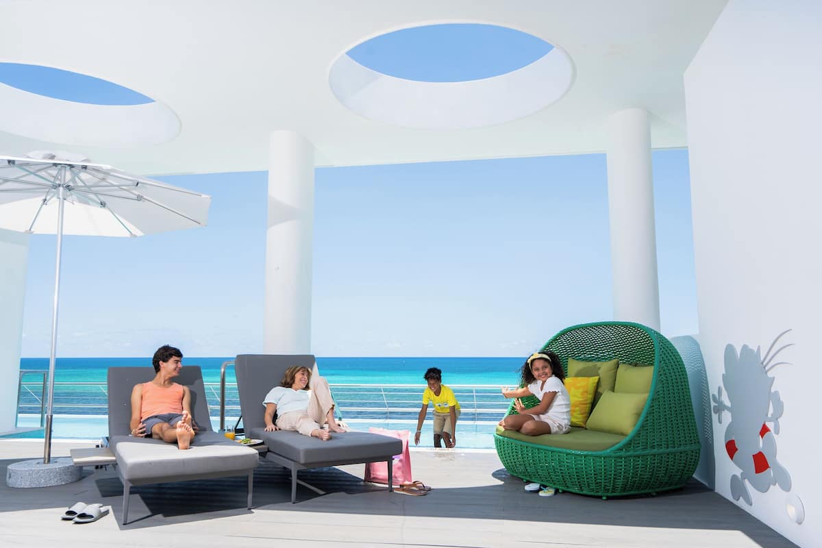 Un espace de détente extérieur moderne, doté de différents types de sièges, notamment un fauteuil en osier vert et des chaises longues, offrant une vue imprenable sur un océan d’un bleu profond.