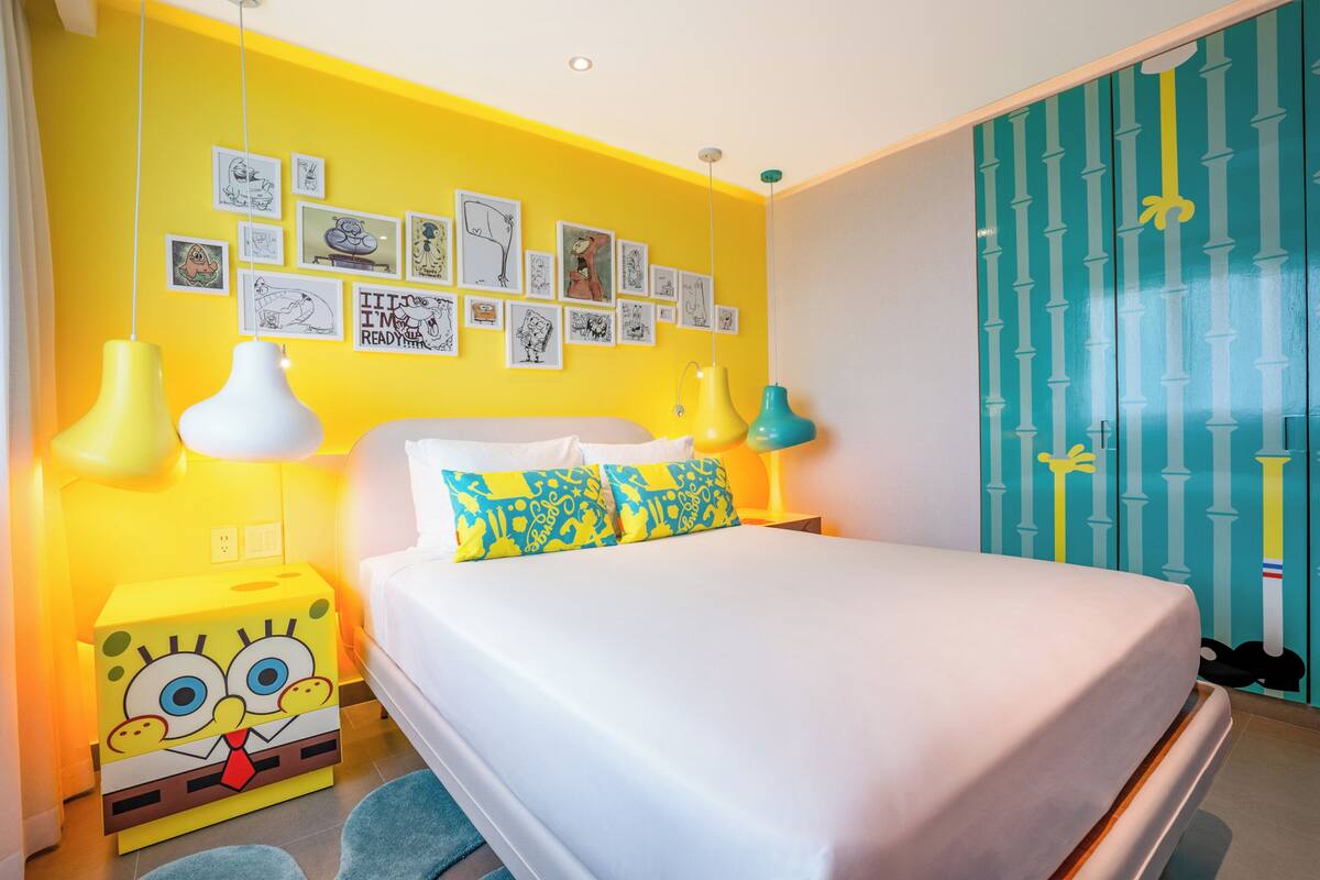 Une chambre aux couleurs vives, avec un lit, des œuvres d’art accrochées au mur et une table de chevet sur le thème de Bob l’éponge.