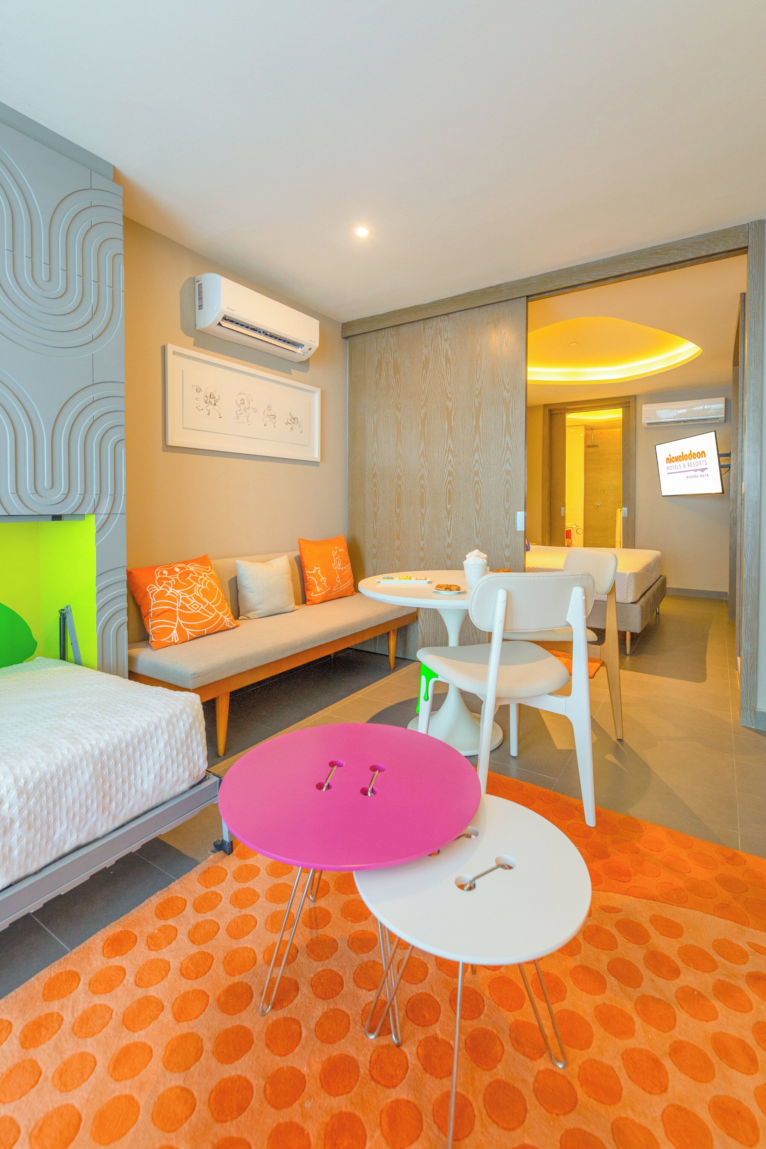 A modern hotel room with a sofa, a small dining table, a pink table, and a bed.