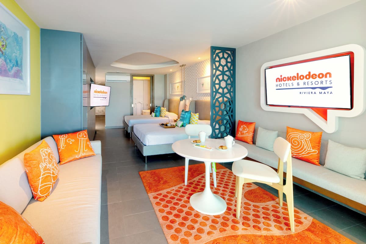 Une chambre d’hôtel comprenant un lit, un canapé, une table à manger et un téléviseur affichant le logo de Nickelodeon Hotels & Resorts.