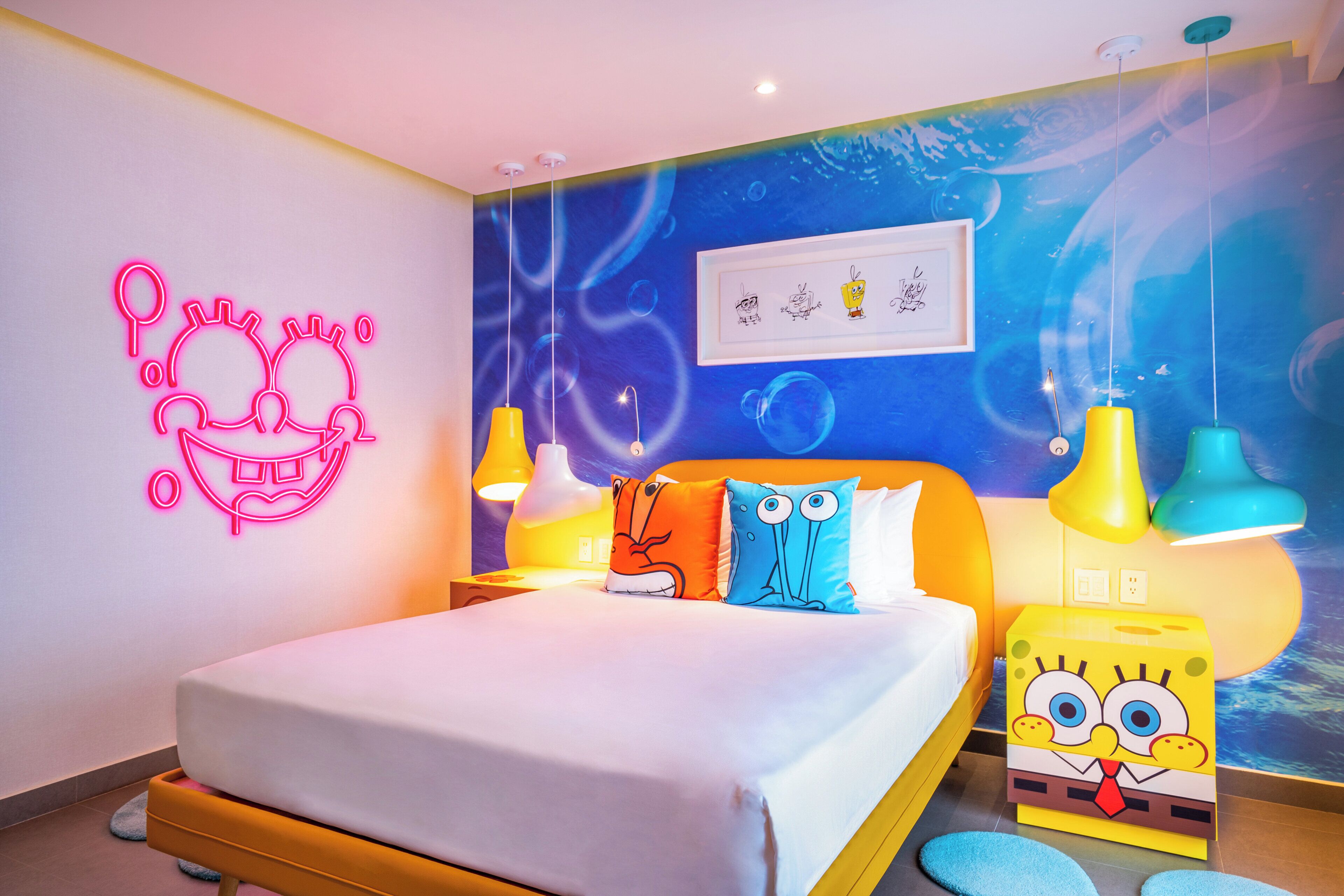 A brightly lit hotel room with a bed, colorful pillows, a neon wall decoration, and a SpongeBob SquarePants-themed nightstand.