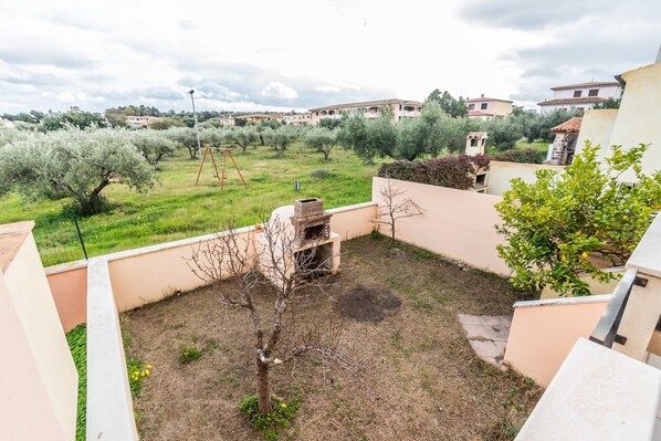 A courtyard with a barbecue area, a small tree, and a view of olive trees and buildings in the background. - Casa Marta (orosei)