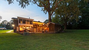 Une maison moderne à deux étages, dotée d’un espace extérieur aménagé et abrité, d’un garage et d’un grand arbre dans le jardin avant.