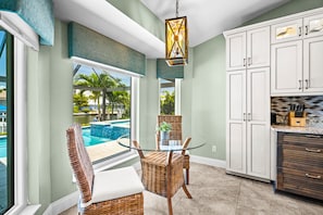 A kitchen with white cabinets, a glass table, wicker chairs, and a view of a swimming pool. - Resort-Style Pool & Hot Tub! Outdoor TV, Fire Pit & Bikes for Island Adventures! (Marco Island)