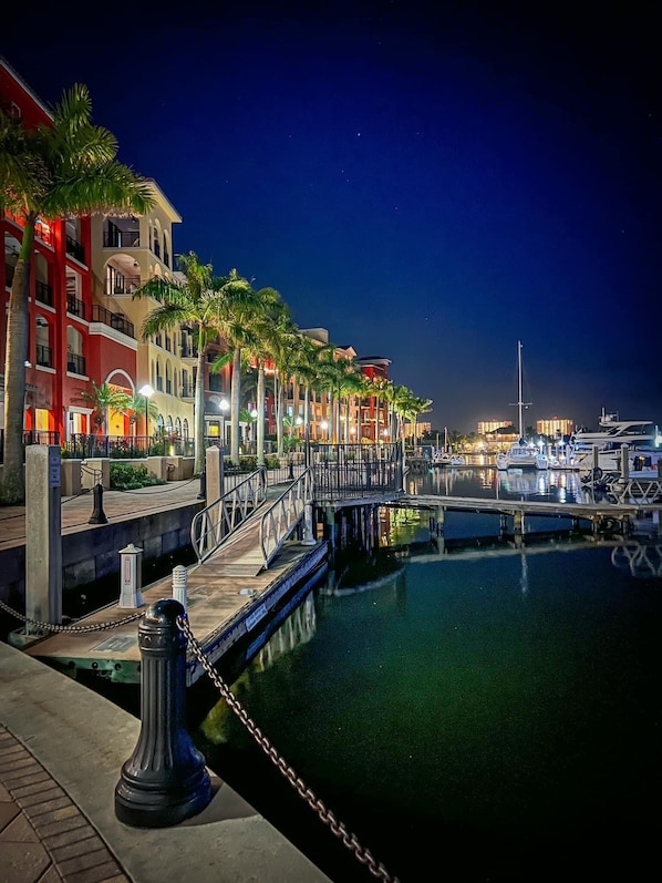 A marina at night with a dock, boats, and a row of buildings with balconies and palm trees. - Resort-Style Pool & Hot Tub! Outdoor TV, Fire Pit & Bikes for Island Adventures! (Marco Island)