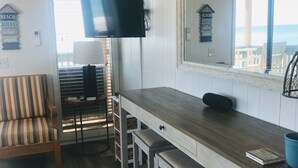 A room with a wooden floor, a striped chair, a bar area with stools, a television mounted on the wall, and a view of the ocean through a window. - Recently Renovated OCEANFRONT CONDO Directly on Beach w/2 bedrooms and 2 baths. (Myrtle Beach)