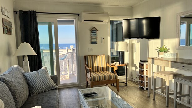 A living room with a sofa, a glass coffee table, a striped armchair, a television, and a view of the beach. - Recently Renovated OCEANFRONT CONDO Directly on Beach w/2 bedrooms and 2 baths. (Myrtle Beach)