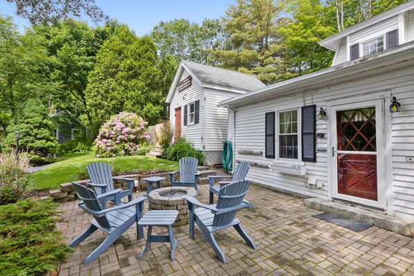 Une terrasse dotée de fauteuils Adirondack, d’un foyer extérieur et d’un chemin en briques menant à une maison blanche avec une porte rouge.