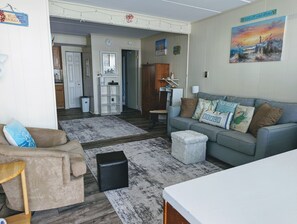 Ein gemütliches Wohnzimmer mit einer grauen Couch, einem beige farbenen Sessel und einem Couchtisch. Im Hintergrund ist eine Küchenzeile erkennbar.
