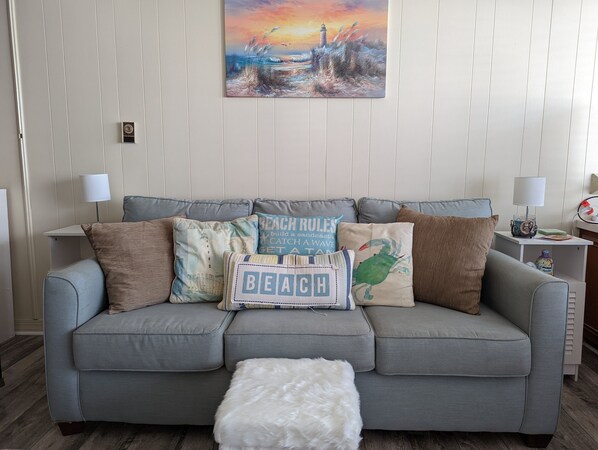 Ein Wohnzimmer mit grauem Sofa, mehreren dekorativen Kissen, einem weißen, flauschigen Hocker und einem Bild einer Strandansicht an der Wand.