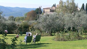 A garden with green chairs, a stone building, and a mountainous backdrop. - Residence La Pieve Panzano in Chianti with Villa Le Barone pool & tennis access  (Panzano In Chianti)