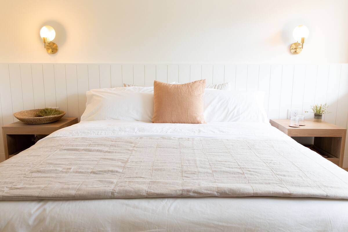 Uma cama bem arrumada com edredom branco, duas almofadas e uma pequena mesa redonda com uma tigela e uma planta.
