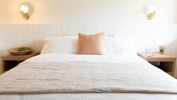 A neatly made bed with a white duvet, two pillows, and a small round table with a bowl and a plant.