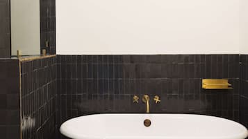 Una bañera blanca independiente con accesorios dorados en un baño con paredes de azulejos negros.
