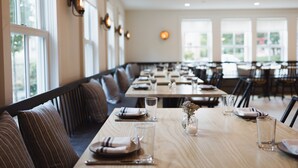 A restaurant with long tables set for dining, benches along the walls, and large windows allowing natural light.