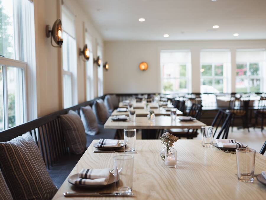 A restaurant with long tables set for dining, benches along the walls, and large windows allowing natural light.