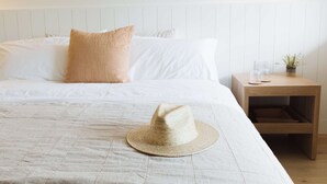 A neatly made bed with white linens and a beige pillow, a straw hat on the bed, a wooden nightstand with a small plant and a glass, and a wall-mounted light fixture.