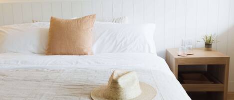 A neatly made bed with white linens and a beige pillow, a straw hat on the bed, a wooden nightstand with a small plant and a glass, and a wall-mounted light fixture.