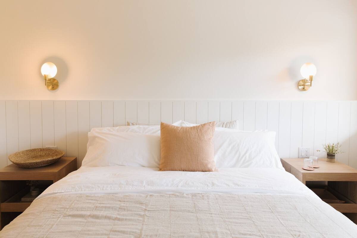 Cama bem arrumada com lençóis brancos e um único travesseiro bege, com duas mesas de cabeceira, uma cesta de vime e um pequeno vaso com planta.