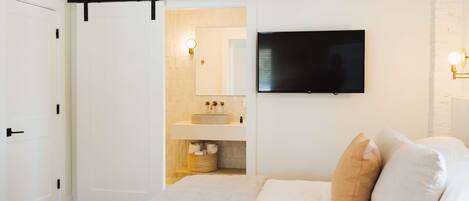 Une chambre à coucher avec un lit, un téléviseur fixé au mur, une porte coulissante donnant sur une salle de bain et un plancher en bois.