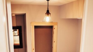 Ein schmaler Flur mit Holzboden und einer Treppe, die zu einer Zwischenebene führt. Eine Leuchte hängt von der Decke, und ein kleiner Tisch mit verschiedenen Gegenständen steht auf der Zwischenebene.
