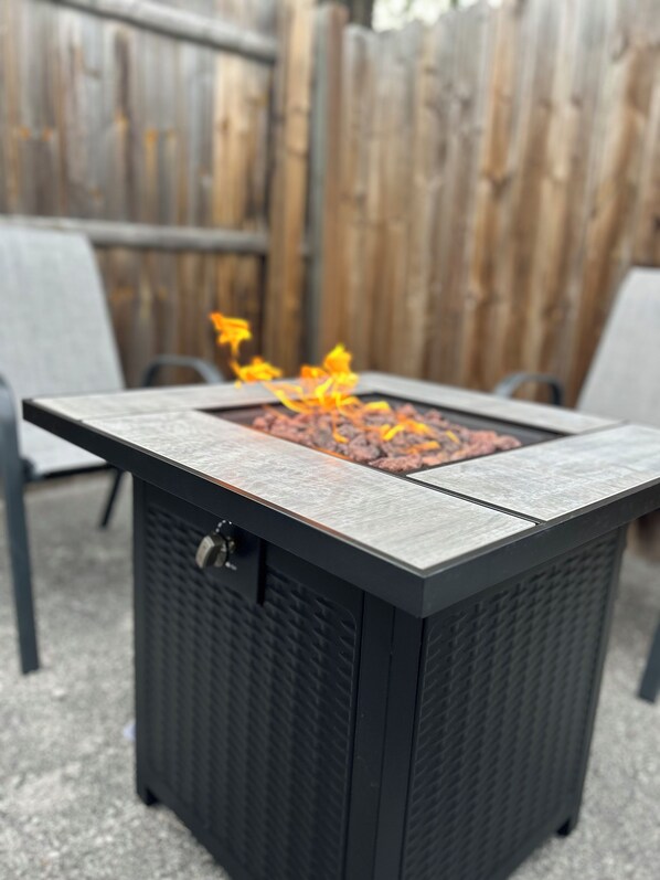 A square outdoor fire feature with a textured top, surrounded by chairs, set against a wooden fence.