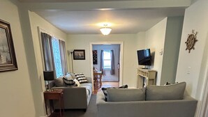 Ein Wohnzimmer mit grauem Sofa, Holzboden, einer Essecke mit Tisch und Stühlen sowie einem Fernseher auf einem Schrank.