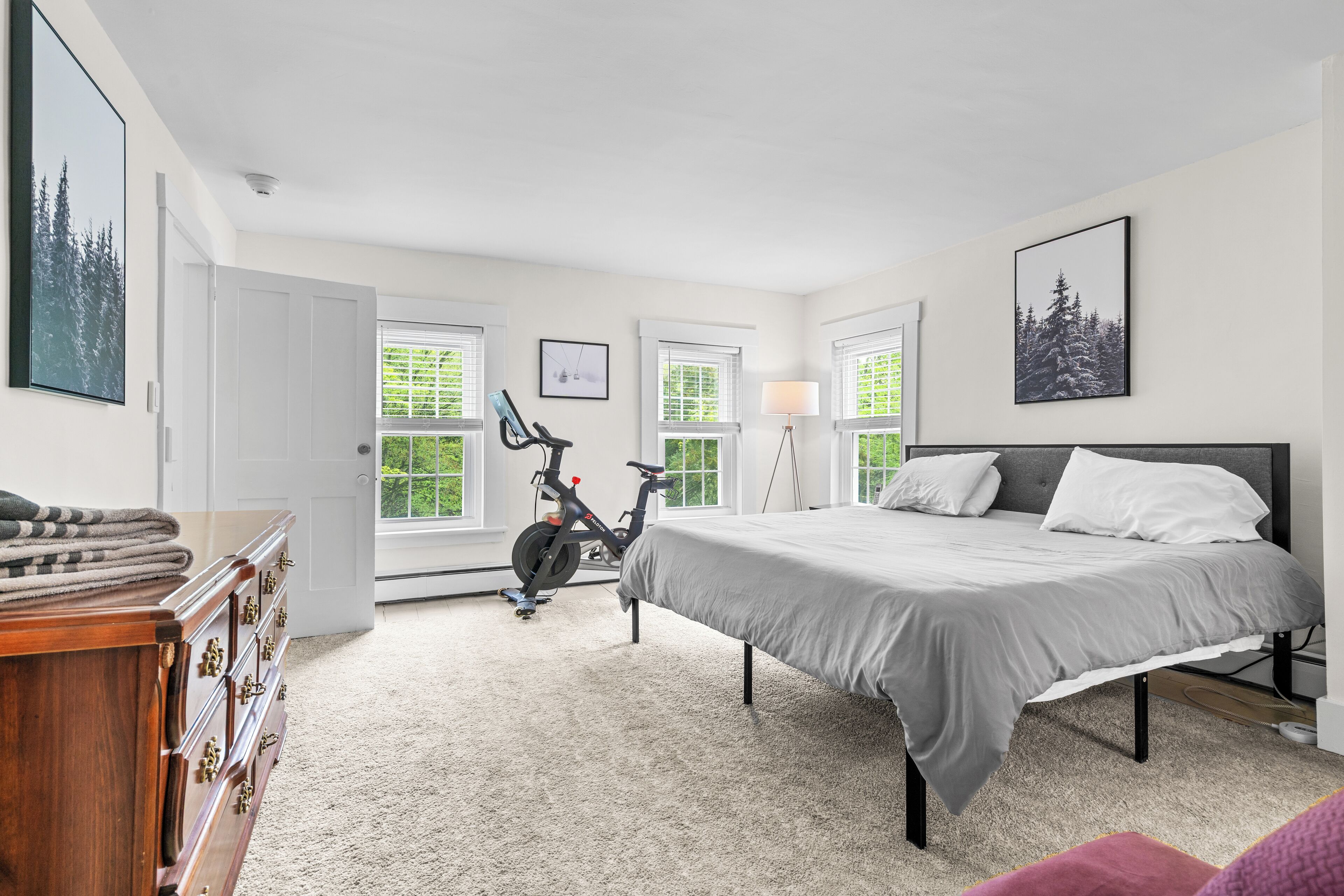 A bedroom with a bed, dresser, exercise bike, and large windows.