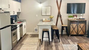 Una cocina compacta con gabinetes blancos, encimera de granito y estufa integrada. Hay una isla de madera con taburetes, un televisor sobre un mueble de madera y un soporte para esquíes en la pared.