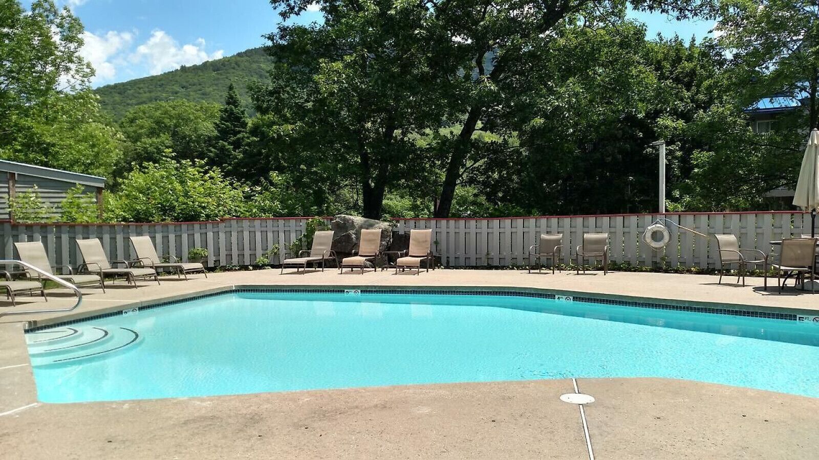 Una piscina rectangular con borde enlosetado, rodeada de sillas de descanso y una cerca blanca.