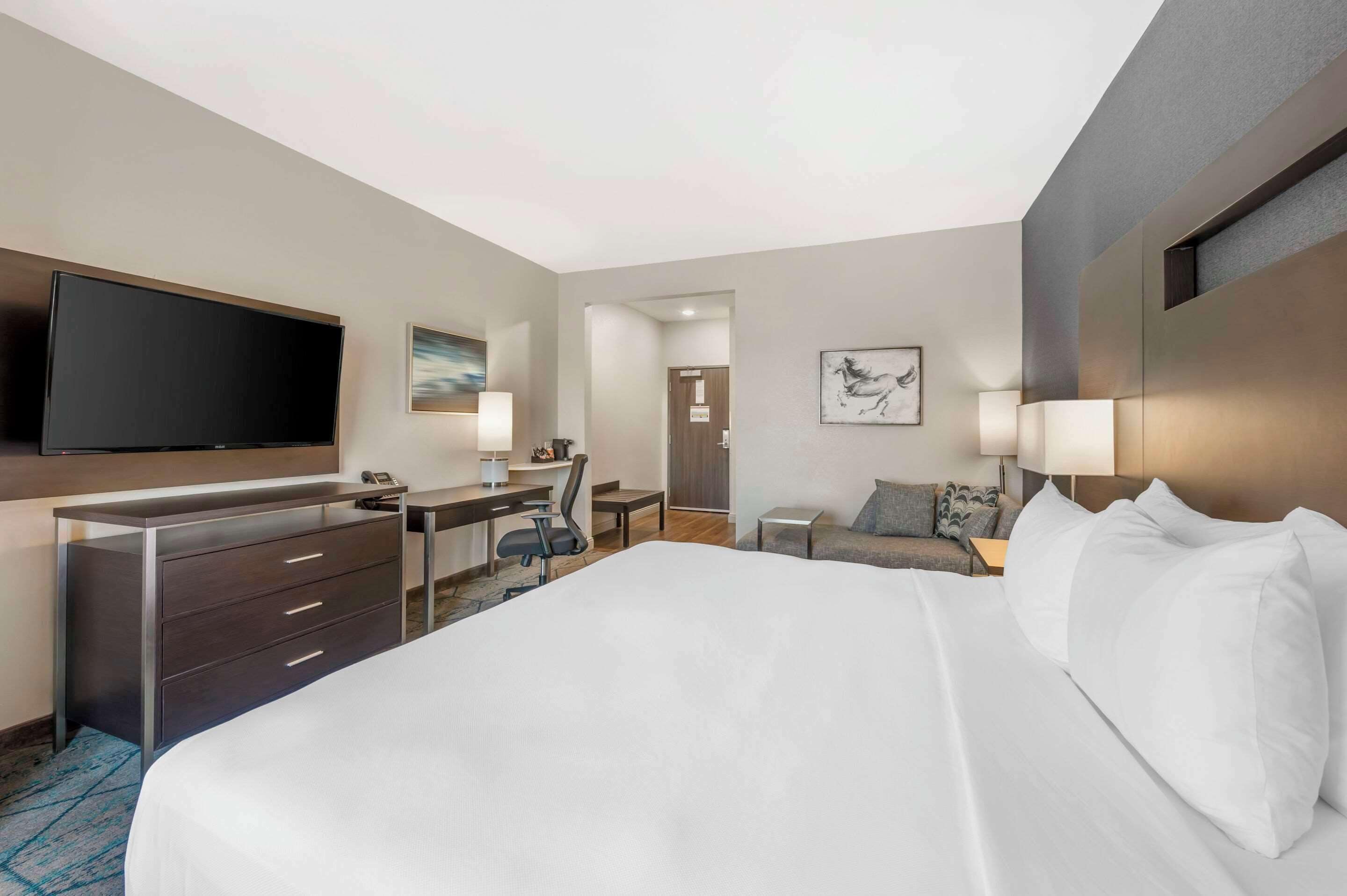 A hotel room with a large bed, a desk with a chair, a flat-screen TV, and a small table with a lamp.