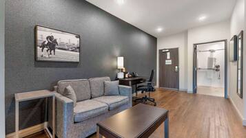 Quarto de hotel moderno com um sofá cinzento, uma mesa pequena, uma escrivaninha com uma cadeira e um quadro emoldurado na parede.