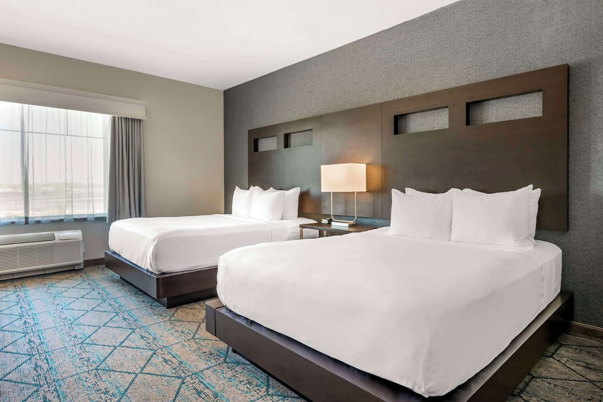 Habitación de hotel con dos camas, un ventanal con cortinas, una lámpara de pared y una alfombra estampada.